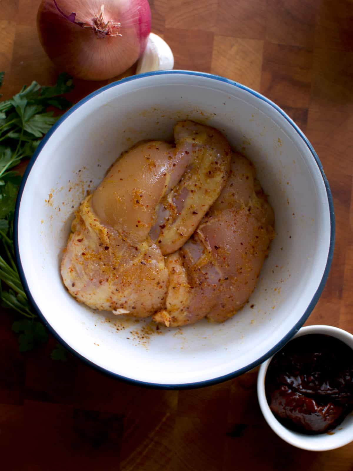 Chicken in a bowl with seasoning, an onion and a small bowl with poblano peppers next to it on a wooden surface.