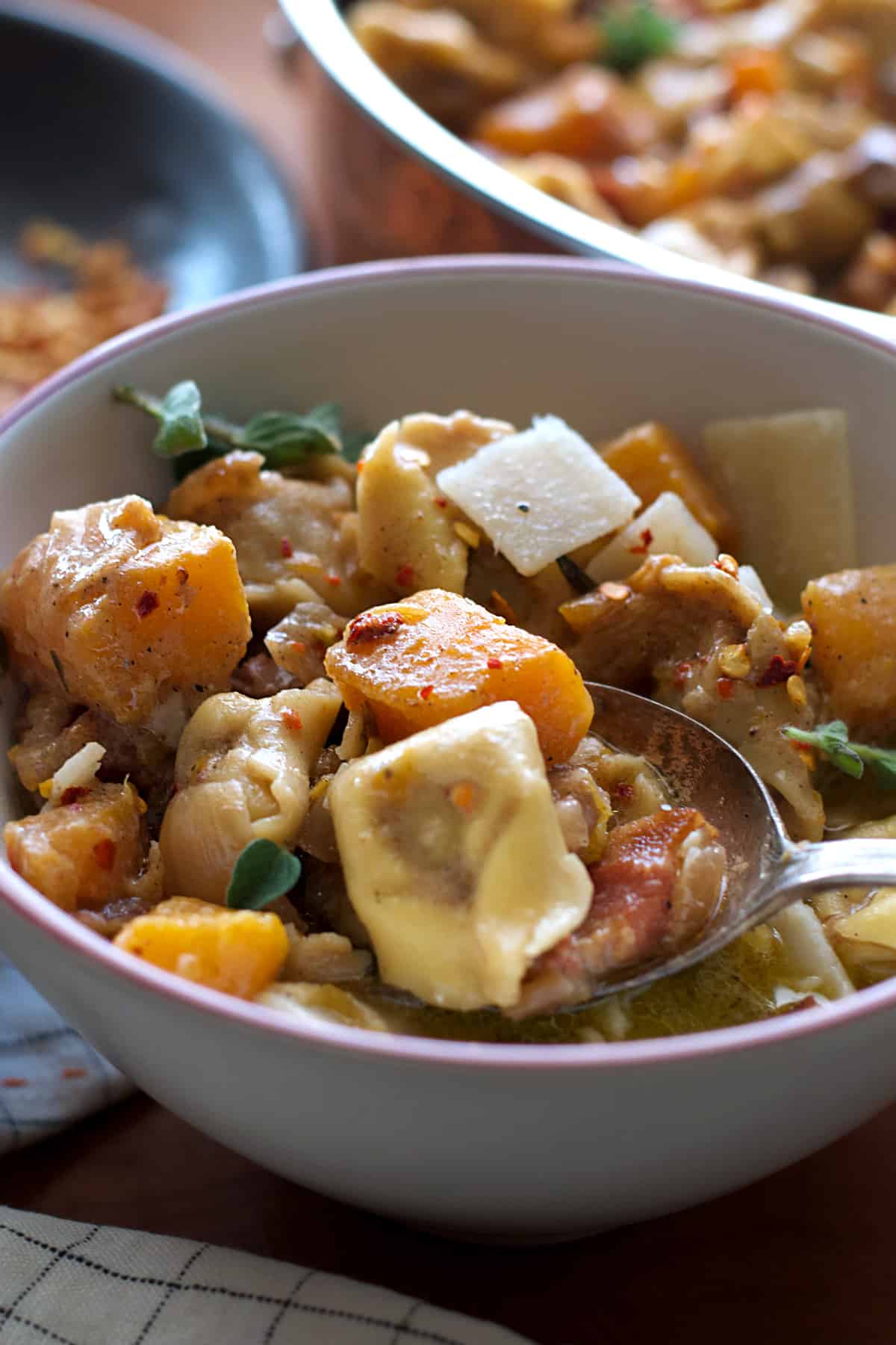 A bowl with pumpkin stew with tortellini and a spoon, half view of a pan with stew and a plate with herbs and spices.