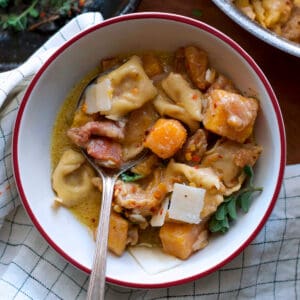 A bowl with pumpkin stew with tortellini and a spoon, partial view of a pan with stew and a plate with herbs and spices.