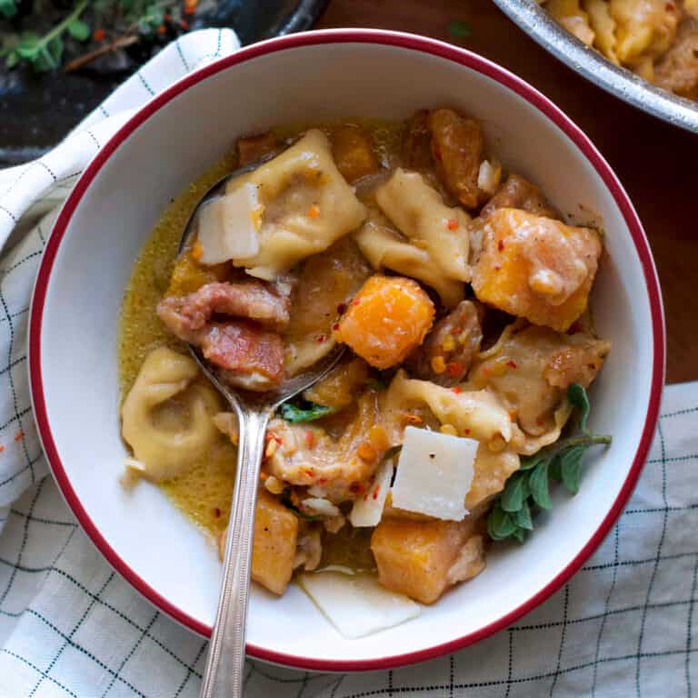 A bowl with pumpkin stew with tortellini and a spoon, partial view of a pan with stew and a plate with herbs and spices.