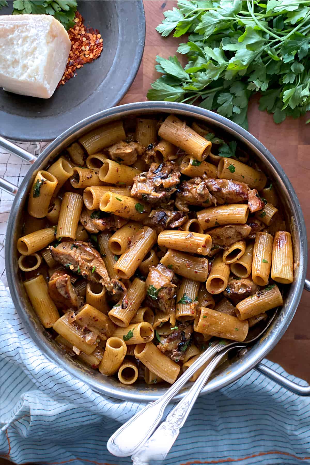 A large deep saucepan with spicy chicken chipotle pasta, two serving utensils. A piece of parmesan cheese and parsley bunch above it. All on a wooden surface.