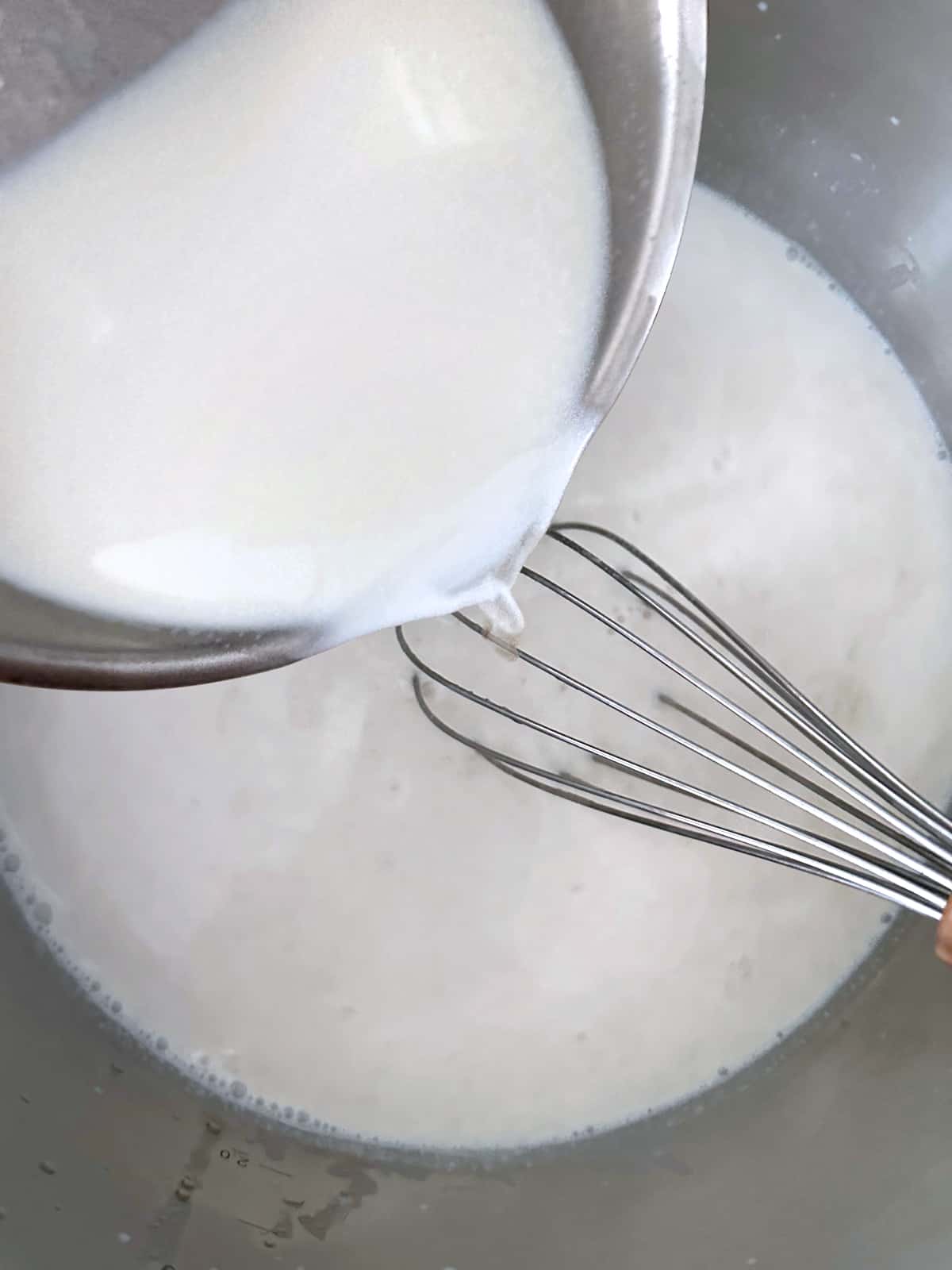 A saucepan with milk poured into white sauce and a whisk.