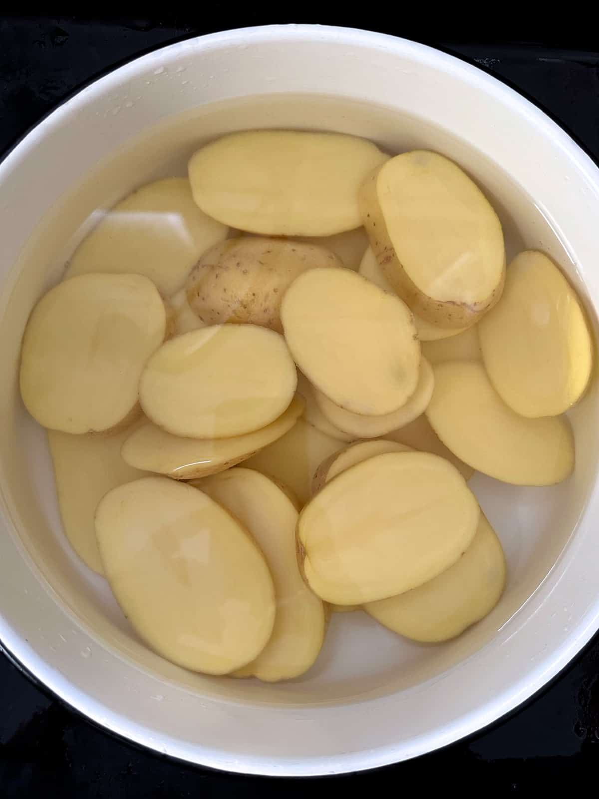 A white bowl filled with sliced potatoes and water in a sink.