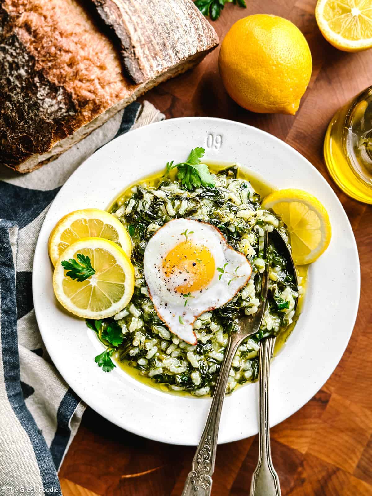 A plate with spanakorizo greek rice with spinach and lemon with a fried egg on top and a fork and spoon. Partial view of a loaf of bread, a lemon and a bottle of olive oil.