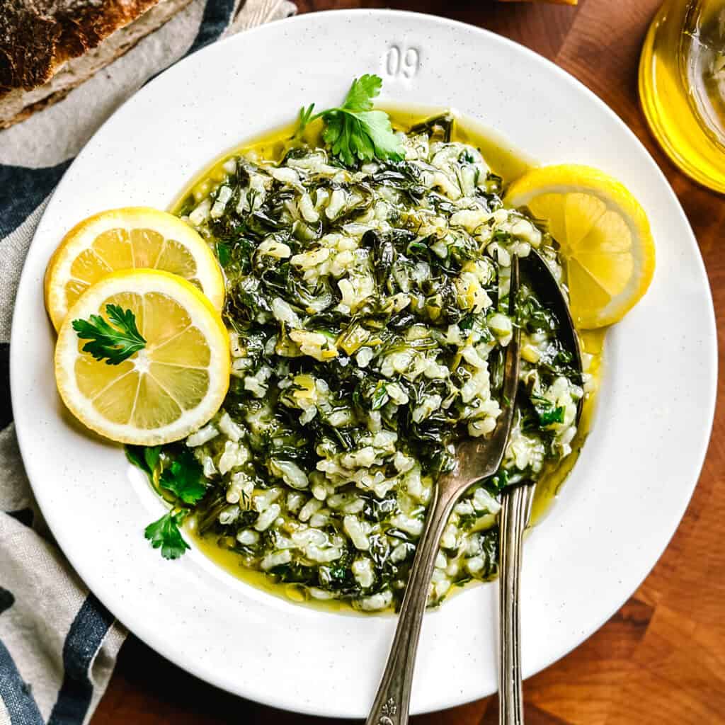 A plate with spanakorizo-Greek rice with spinach and lemon, a couple of lemon slices and a fork and spoon. Partial view of a loaf of bread, a lemon and a bottle of olive oil.