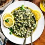 A plate with spanakorizo-Greek rice with spinach and lemon, a couple of lemon slices and a fork and spoon. Partial view of a loaf of bread, a lemon and a bottle of olive oil.