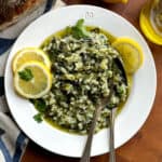 A plate with spanakorizo, a couple of lemon slices and a fork and spoon. Behind it a loaf of bread, a lemon and lemon half and a bottle of olive oil.