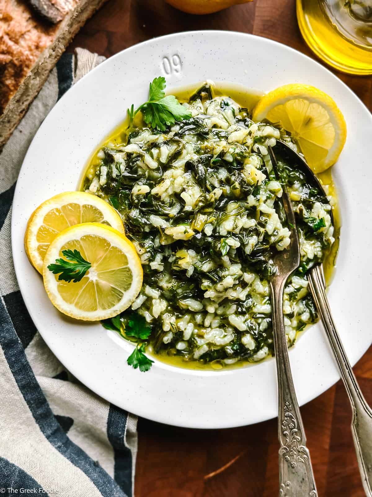 A plate with spanakorizo, a couple of lemon slices and a fork and spoon. Partial view of a loaf of bread, a lemon and a bottle of olive oil.