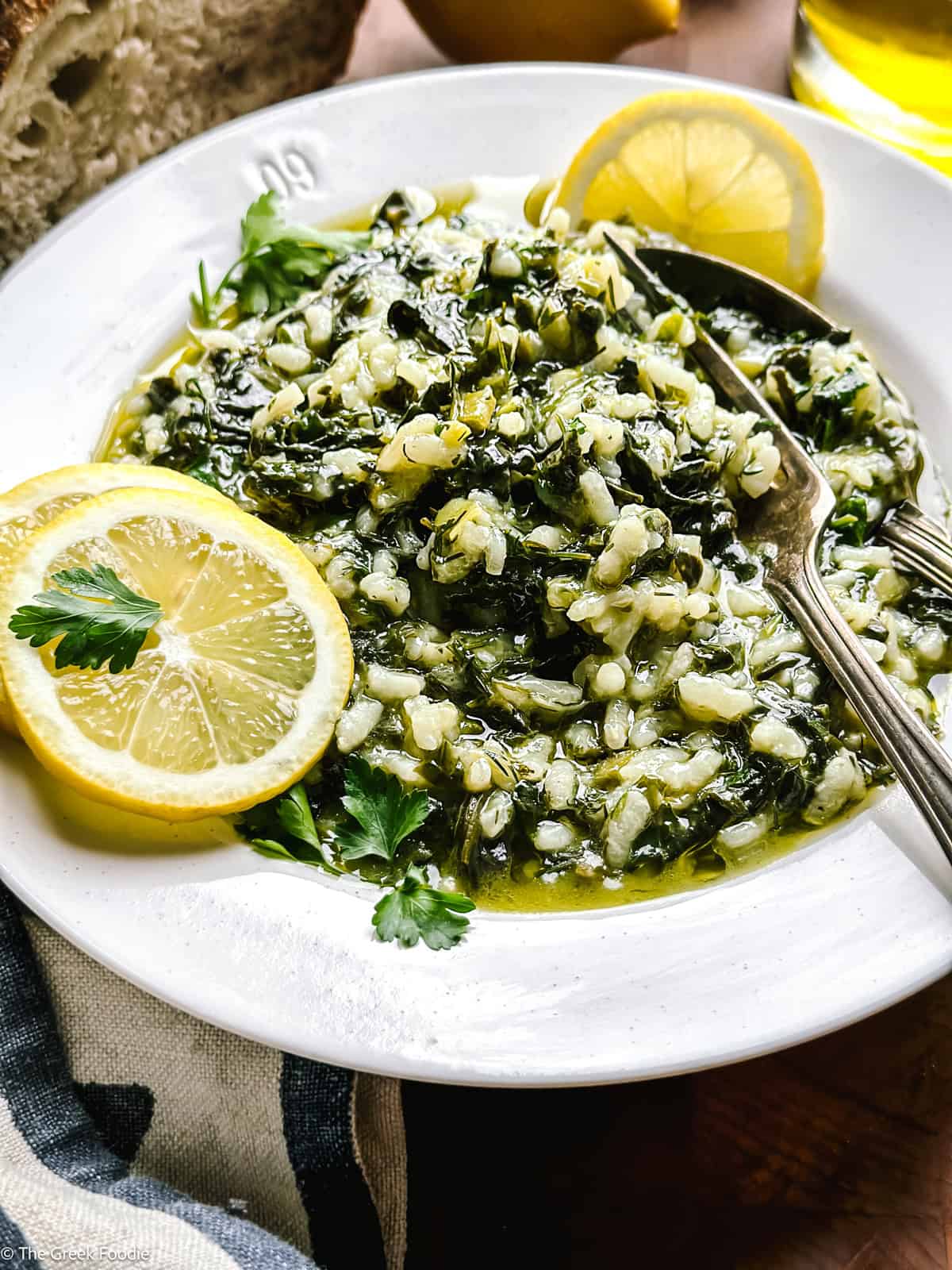 A plate with spanakorizo Greek rice with spinach and lemon, a couple of lemon slices and a fork and spoon. Partial view of a loaf of bread, a lemon and a bottle of olive oil.