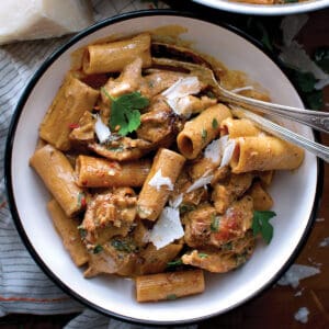 A bowl with Mexican-inspired spicy chicken chipotle pasta, two serving utensils. A piece of parmesan cheese and parsley bunch above it. All on a wooden surface.