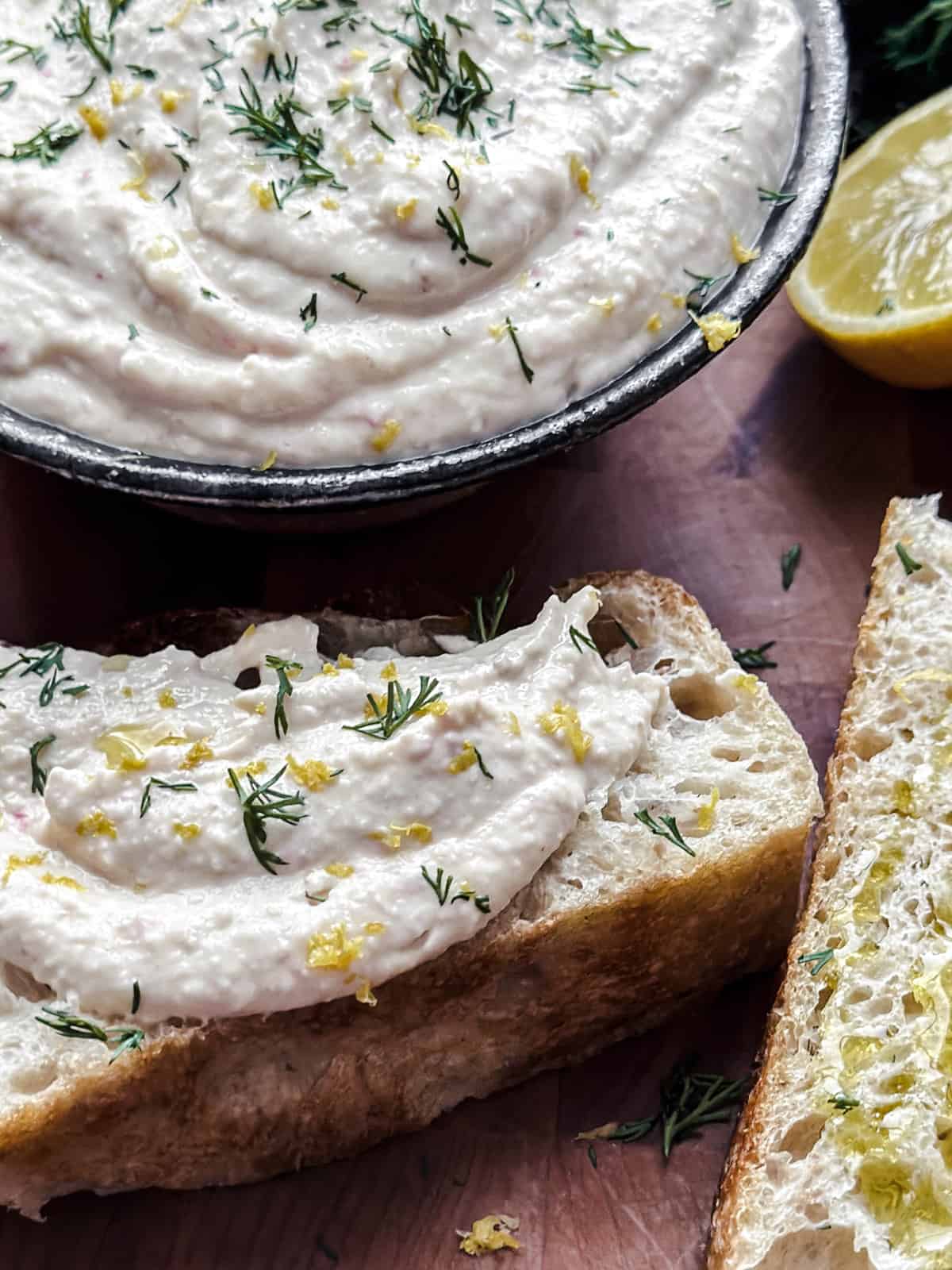 A bowl with taramosalata. Around it pieces of bread with the spread, and a lemon cut in half.