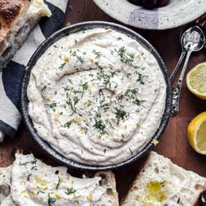 A bowl with taramasalata in the center. Around it pieces of bread, a lemon cut in half, a bowl of olives and two spoons.