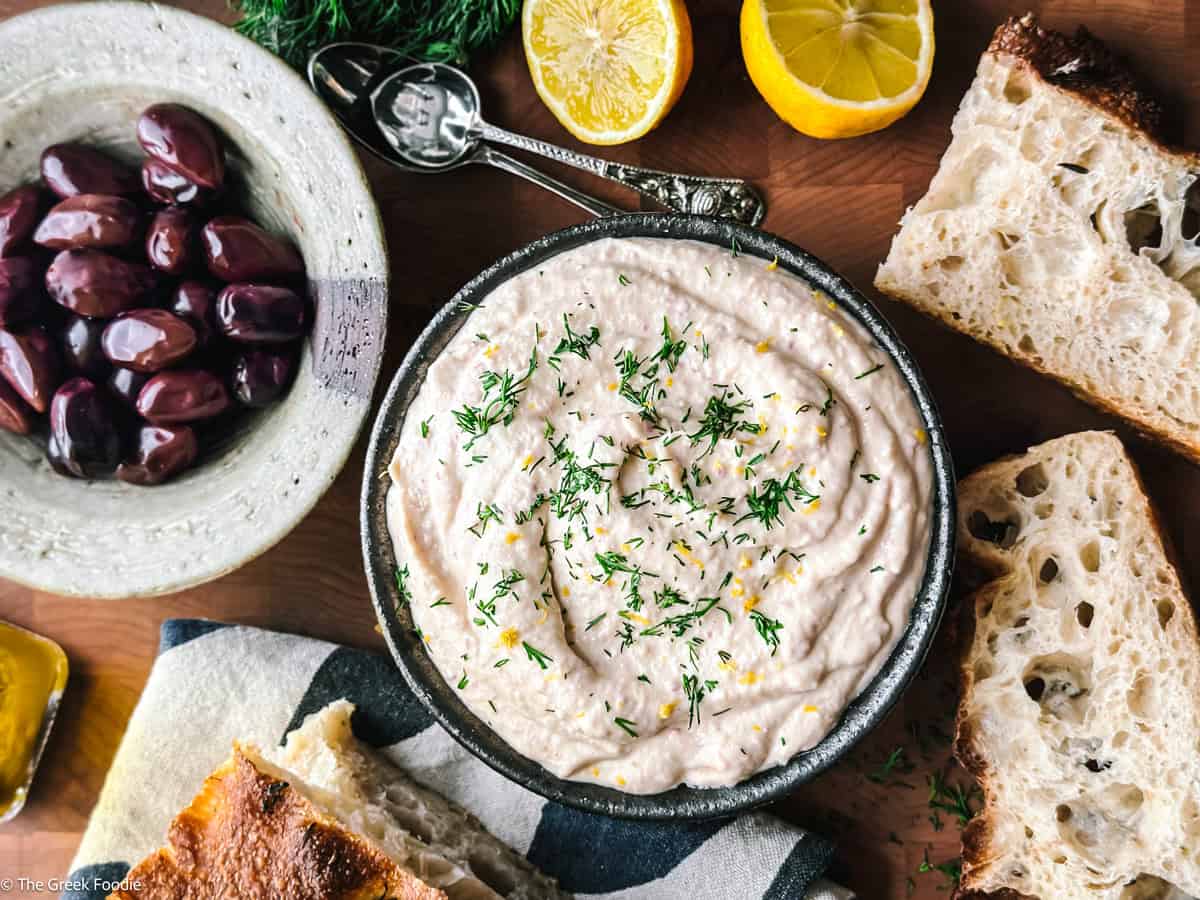 Taramosalata in a white bowl drizzled with olive oil, surrounded by bread, olives, and lemon
