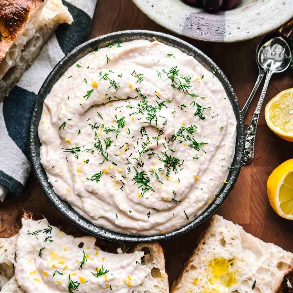 Taramosalata in a white bowl drizzled with olive oil, surrounded by bread, olives, and lemon