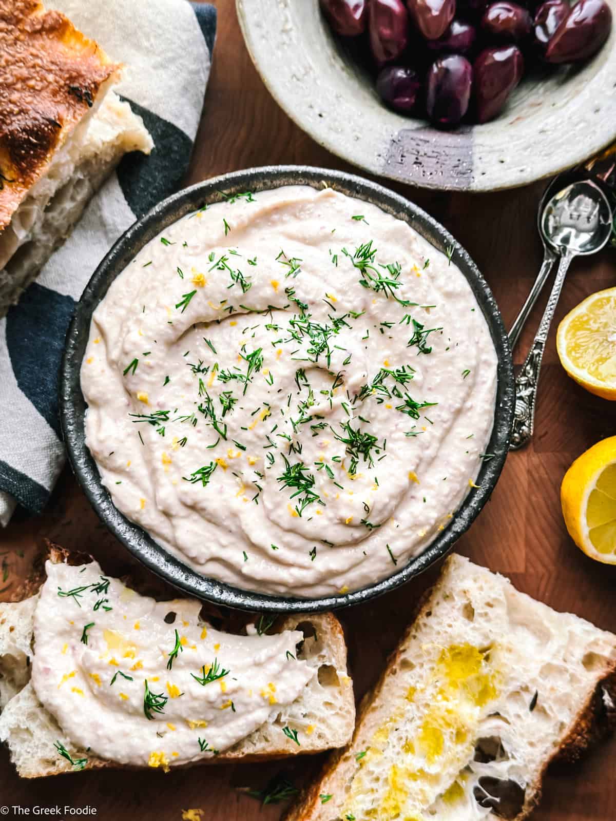 Taramosalata in a white bowl drizzled with olive oil, surrounded by bread, olives, and lemon