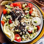 A bowl with two lamb burgers, orzo and tomato salad and lemon wedges.