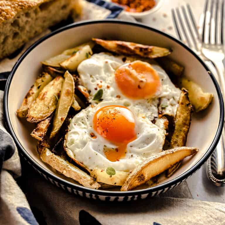 A plate with two fried eggs and oven fries with a loaf of bread behind it, next to utensils and a cloth napkin.