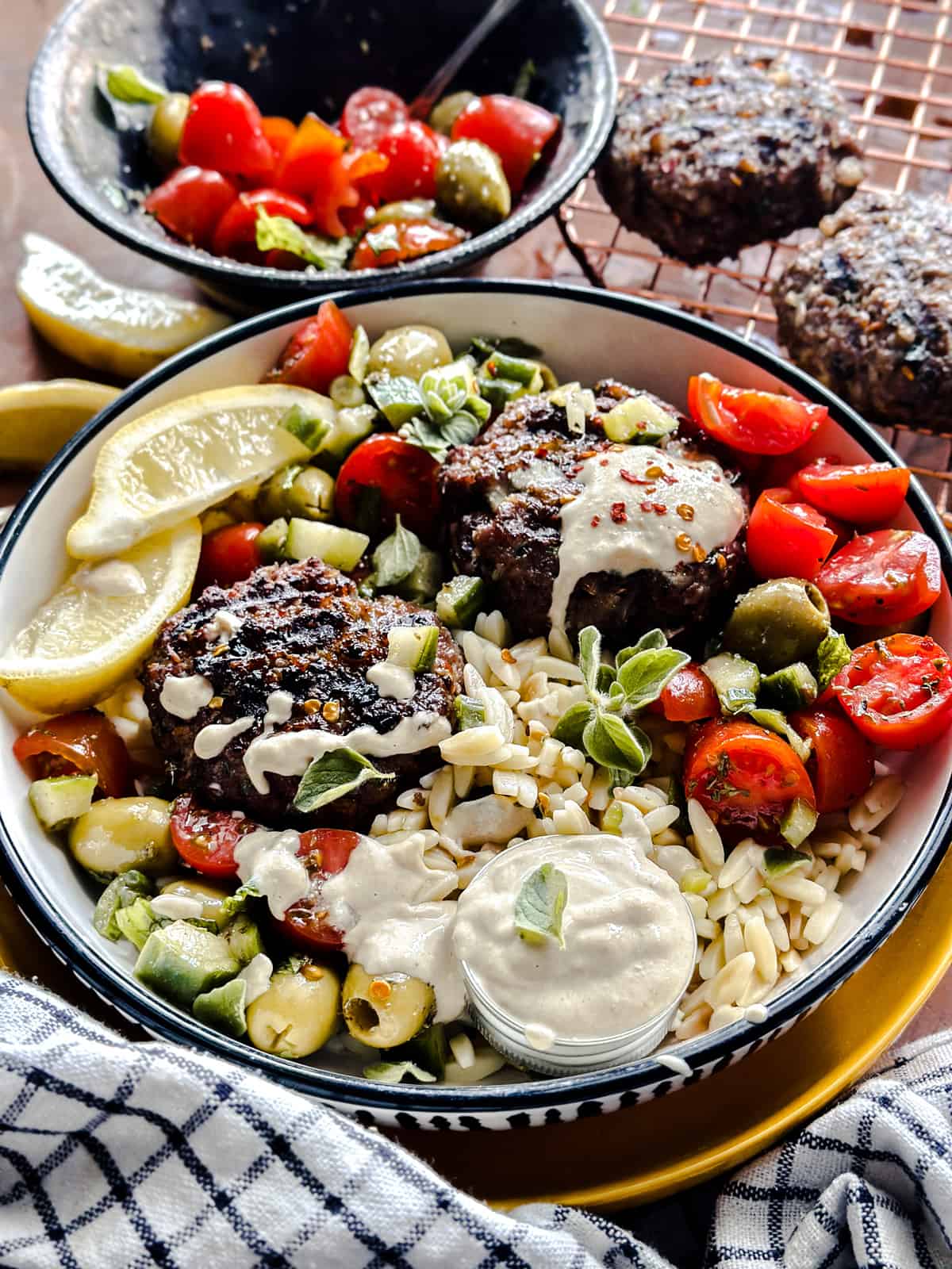 A bowl with two lamb burgers, orzo and tomato salad and lemon wedges. On the table a bowl with salad more lemon wedges and a wire rack with burgers.