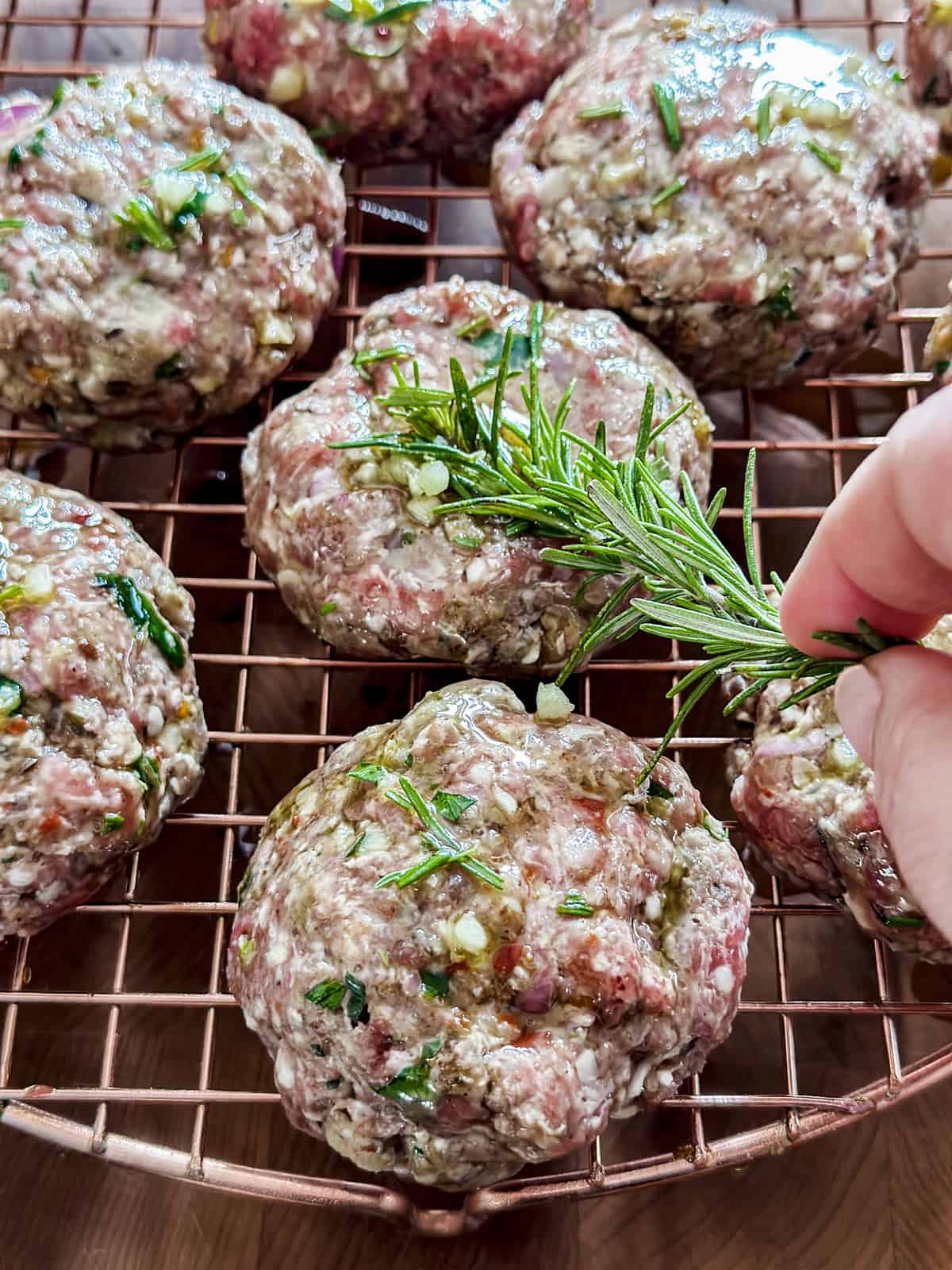 A hand holding a rosemary sprigs brushing olive oil on lamb burgers on a wire rack.