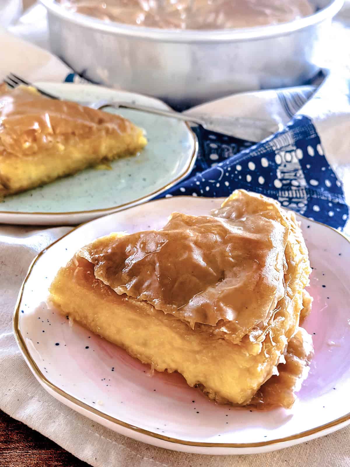 Two pieces of galaktoboureko on plates on top of a cloth napkin, behind them an aluminum pan
