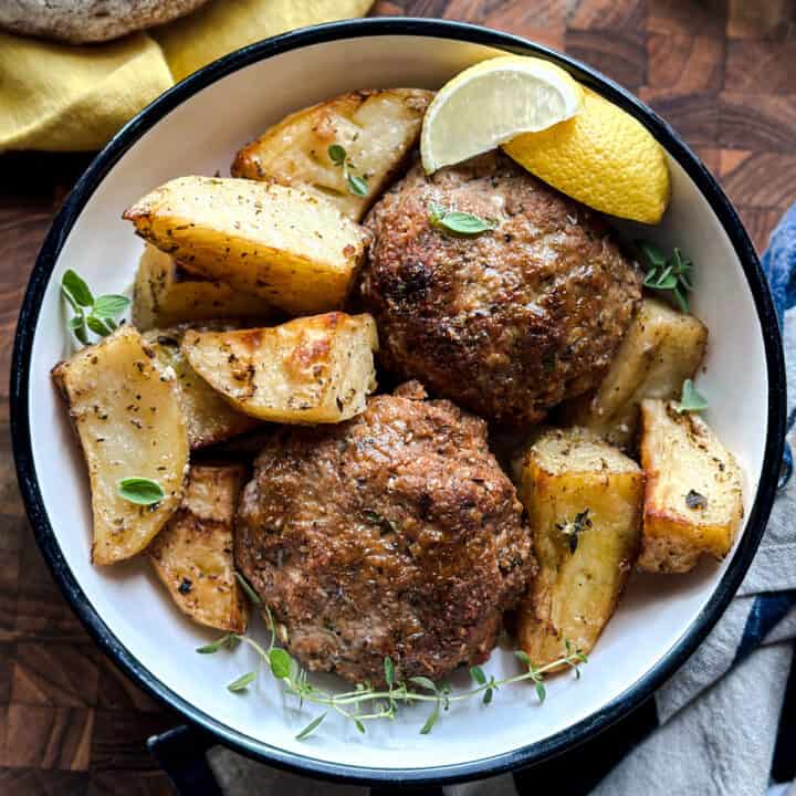 A plate with two biftekia, greek burgers and roasted potatoes on a cloth napkin, next to a loaf of bread on a wooden table.