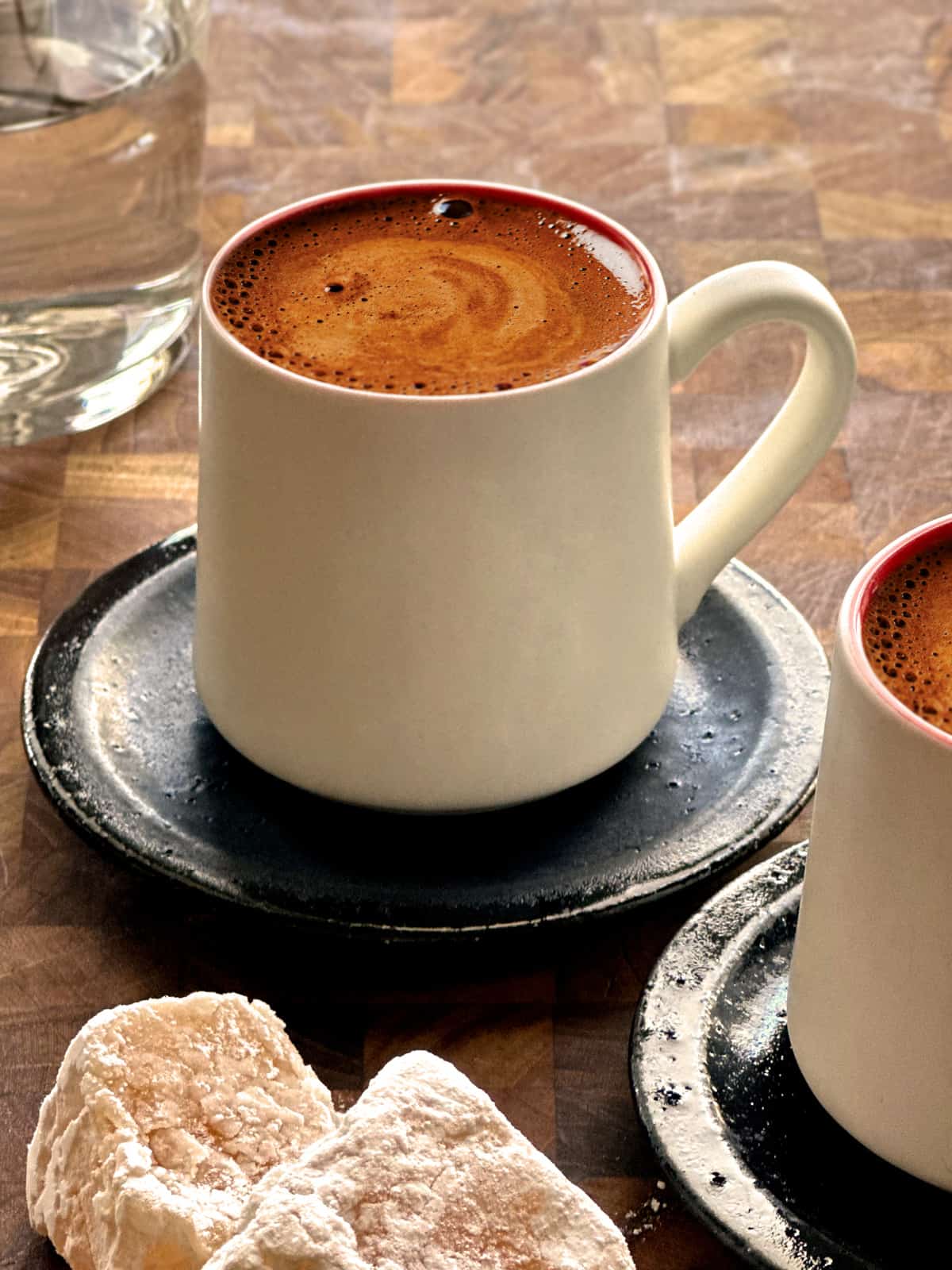 A white cup with Greek coffee on black saucer, and partial views of another cup and a glass of water . At the bottom are two Turkish delights.
