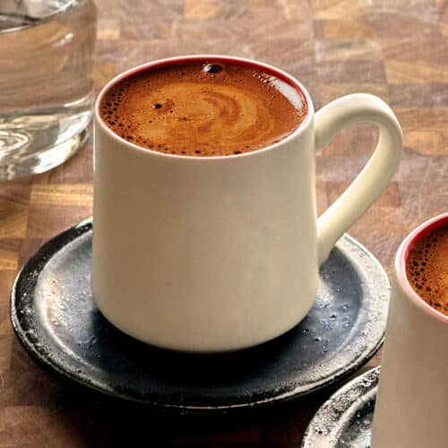 A white cup with Greek coffee on a black saucer, partial view of another cup and a glass of water.