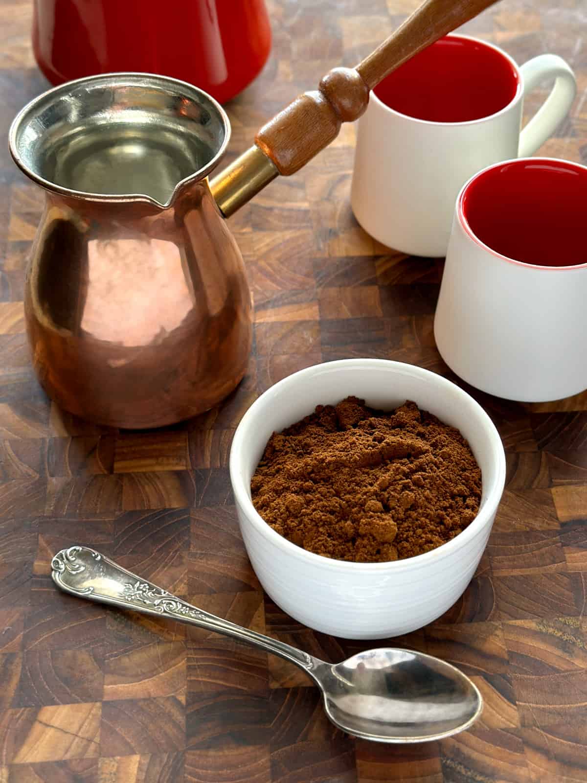 A copper pot, two small coffee cups, a bowl with greek coffee, and a spoon, on a wooden surface.