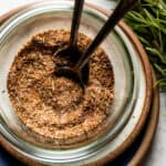 Close up of a bowl with Greek seasoning blend and two spoons. Next to it rosemary leaves.