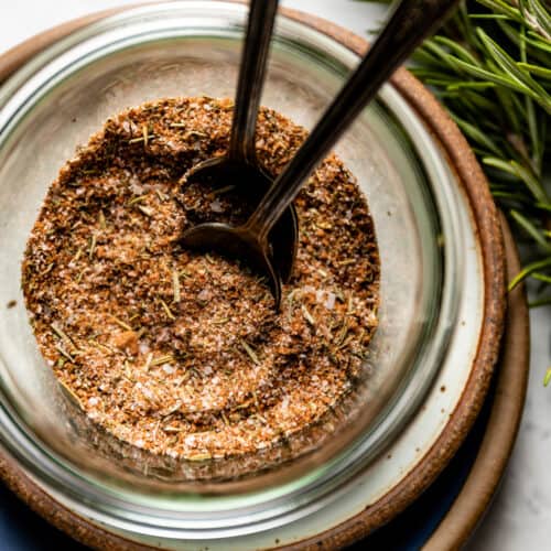 Close up of a bowl with Greek seasoning blend and two spoons. Next to it rosemary leaves.