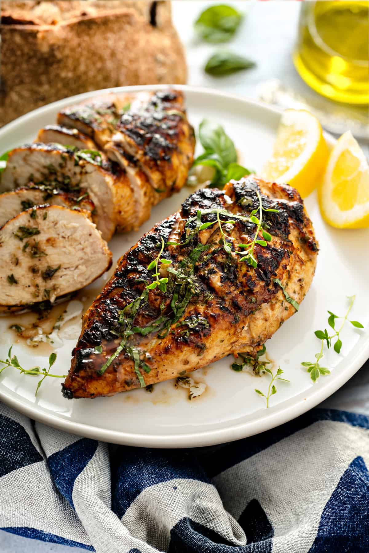 Two chicken breast covered with Greek seasoning on a plate, one of them sliced, two lemon wedges. At the back a loaf of bread and a bottle of olive oil.