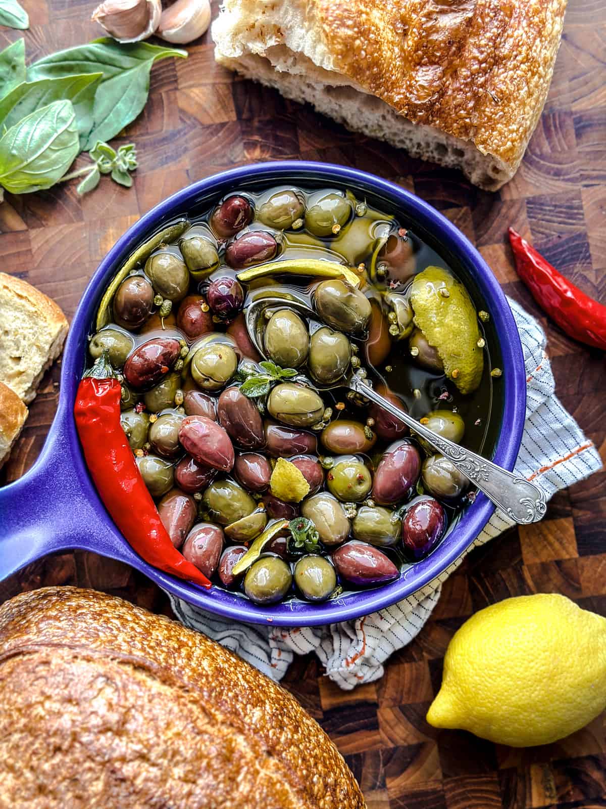 A shallow bowl with marinated olives in olive oil, lemon peel and garlic. A cloth napkin, pieces of bread and herbs are around, on a wooden table.