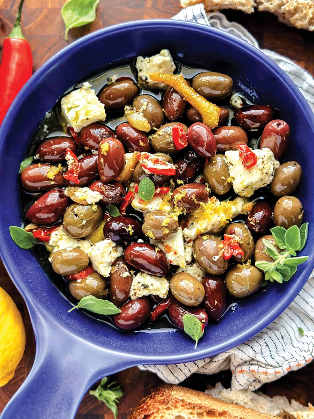 Close up of a shallow bowl with marinated olives in olive oil, lemon peel, feta cheese, and herbs.