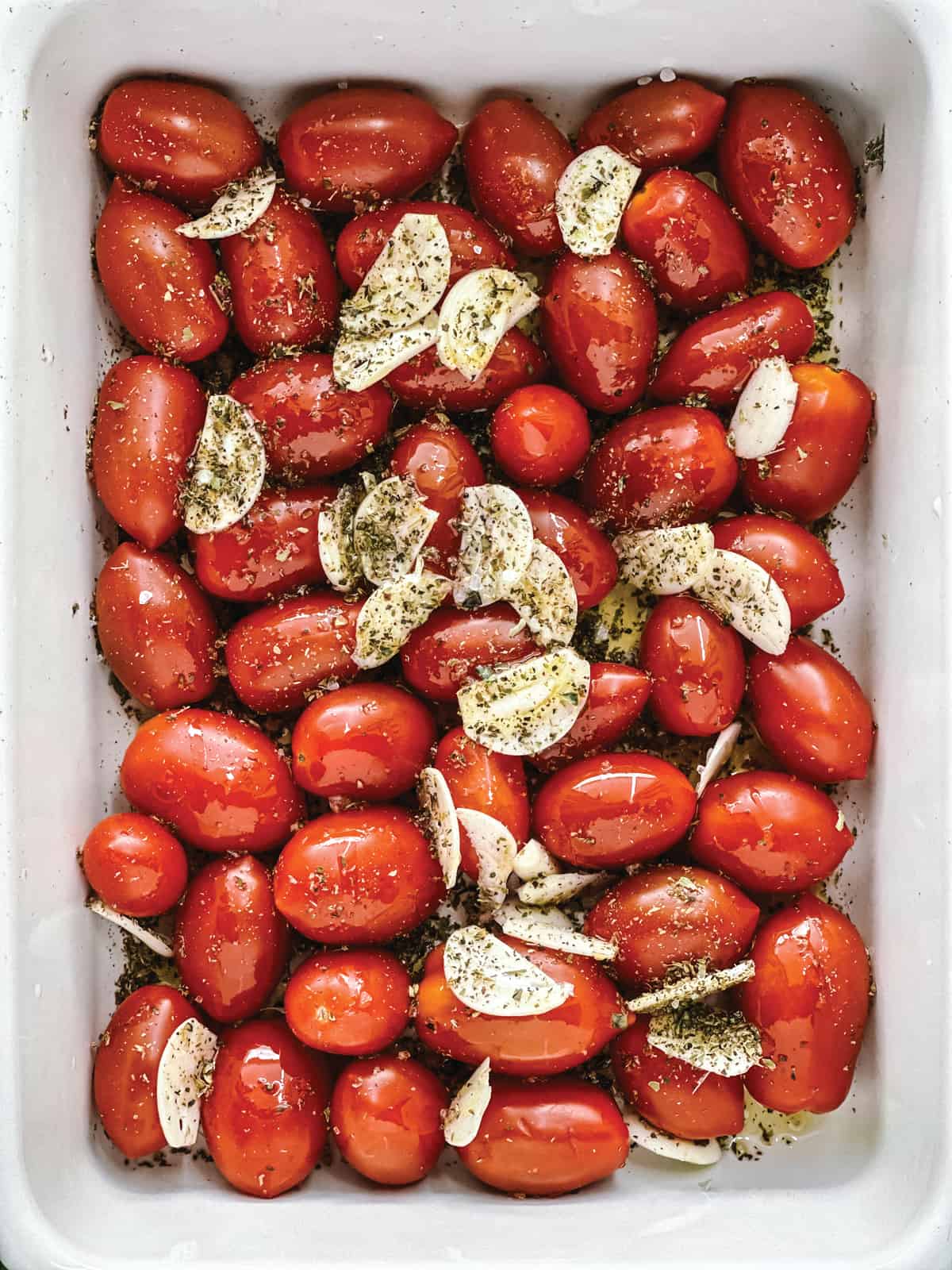 Cherry tomatoes in a white baking pan with garlic slices, olive oil and seasoning.