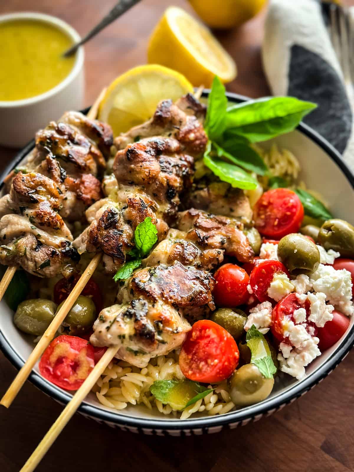A bowl with three chicken skewers orzo pasta , tomatoes and olives, lemons and a container with sauce. 
