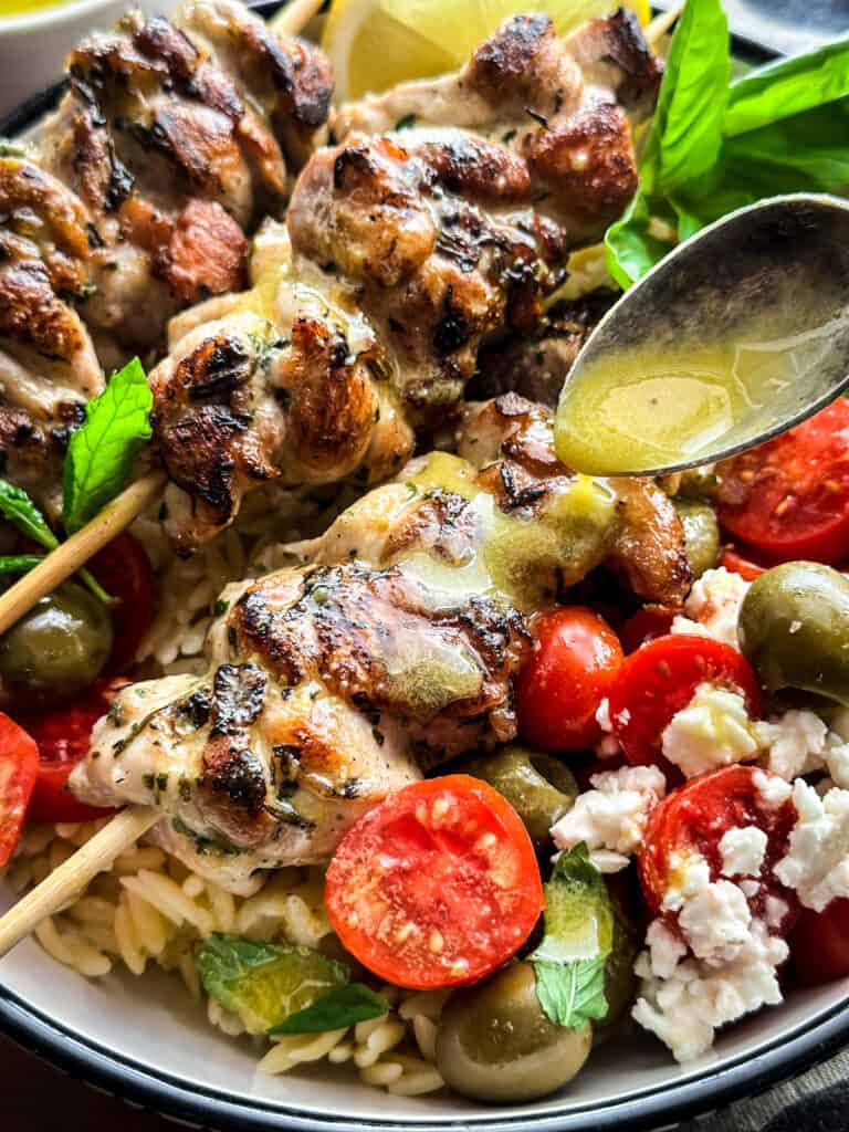 A bowl with three chicken skewers orzo pasta , tomatoes and olives and a spoon pouring lemon sauce.