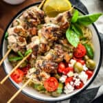 A bowl with three chicken skewers orzo pasta , tomatoes and olives and a lemon wedge.