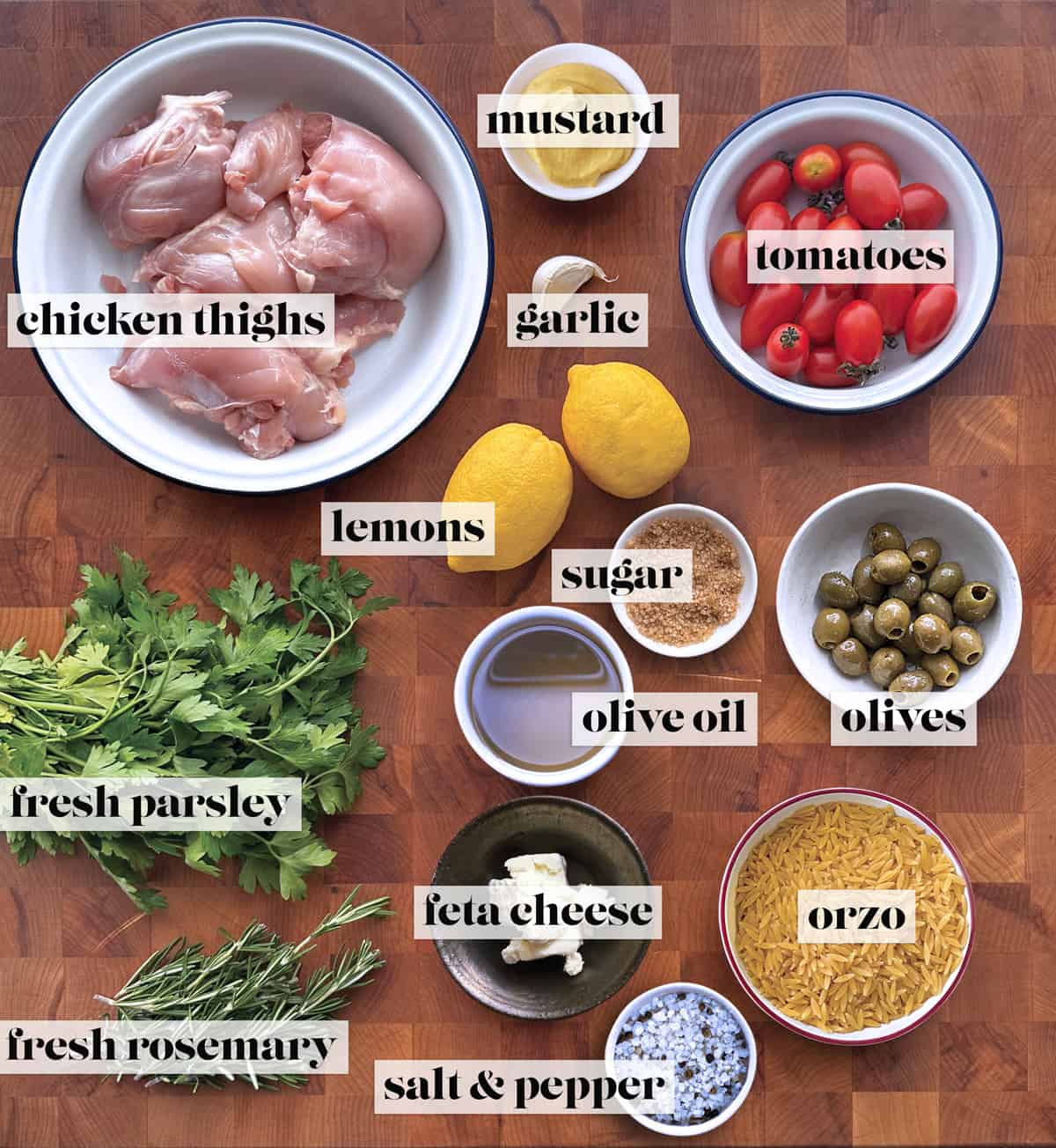 A bowl with chicken things, small bowls with sugar, mustard, sea salt and pepper, orzo pasta, olives, feta cheese, cherry tomatoes, olive oil, two lemons, a clove of garlic, fresh parsley and rosemary on a butcher block.