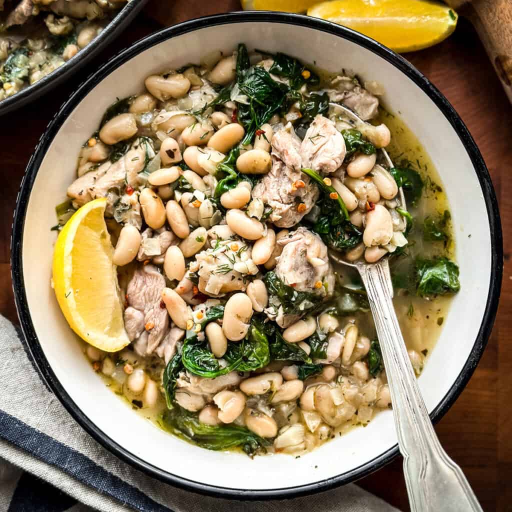 A bowl with chicken stew a spoon and fork, a lemon wedge, a pot with stew at the back and a lemon.