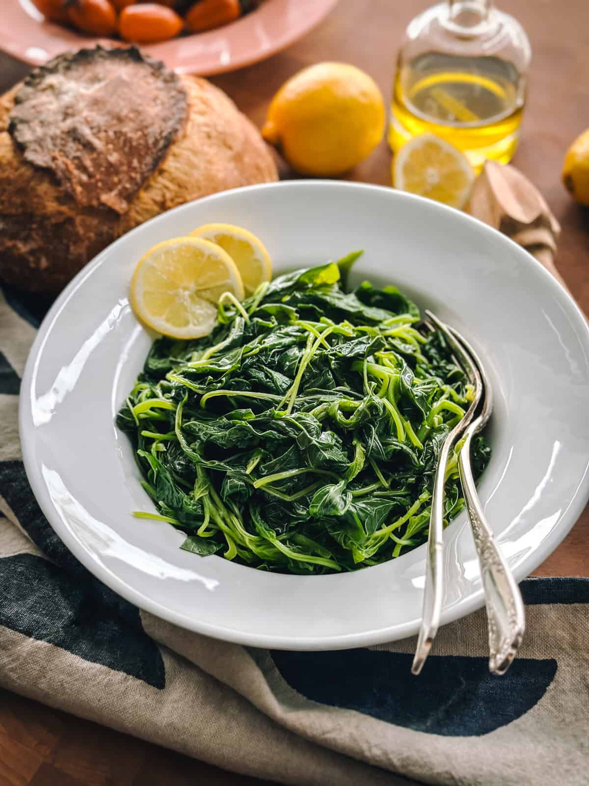 A plate with horta vrasta-boiled Greek greens with two slices of lemon and utensils. A loaf of bread and a cloth napkin, a bottle with olive oil a lemon and lemon halves, all on a table.