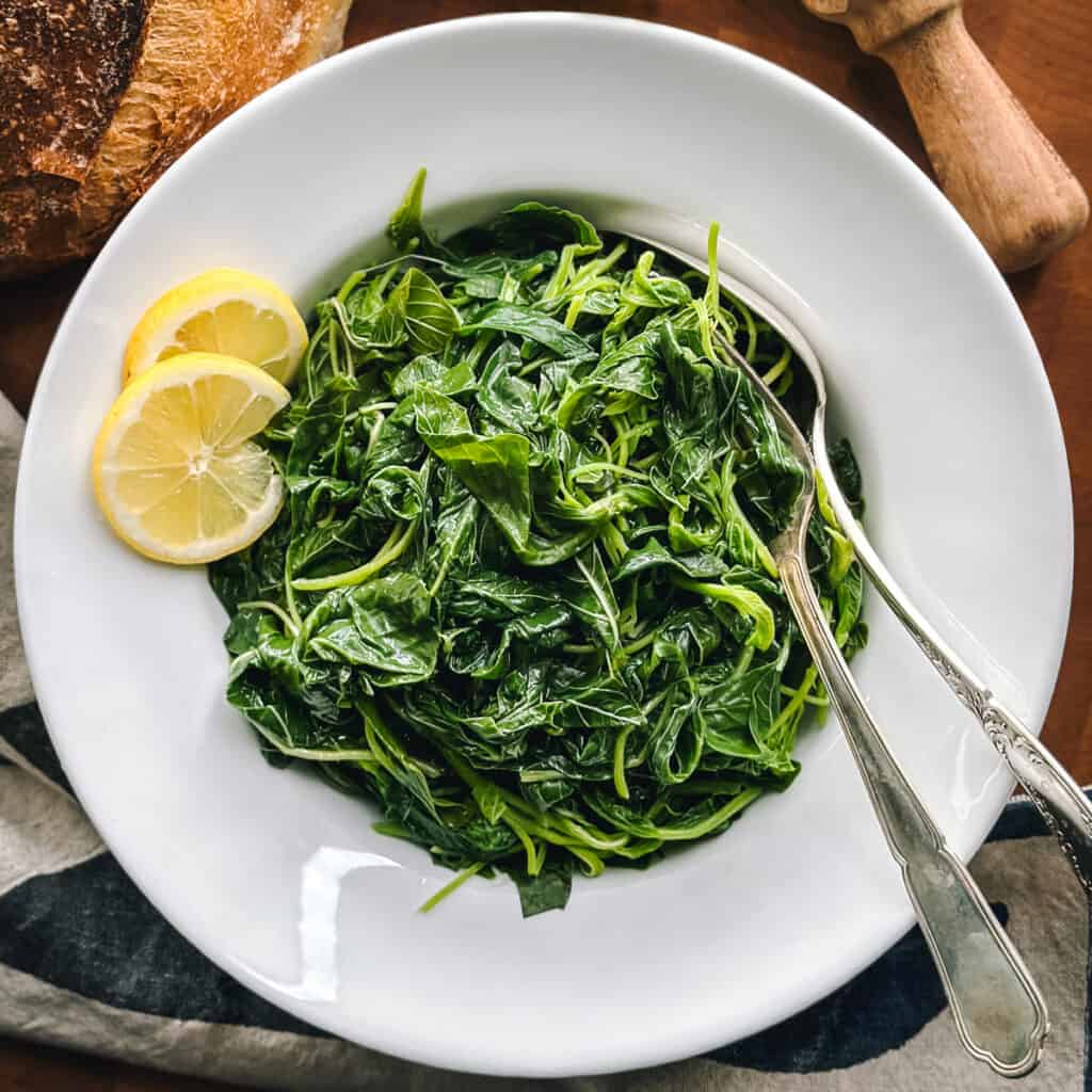 A plate with horta vrasta-Greek greens wit two slices of lemon and utensils.