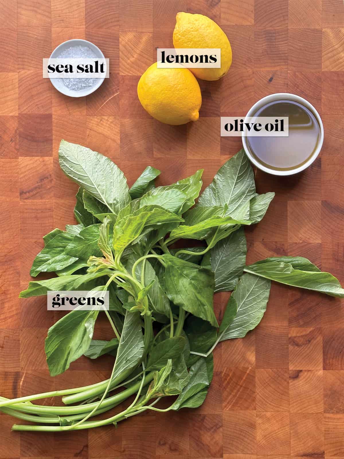 A bunch of greens, two lemons, a bowl with olive oil and a small bowl with sea salt on a table.