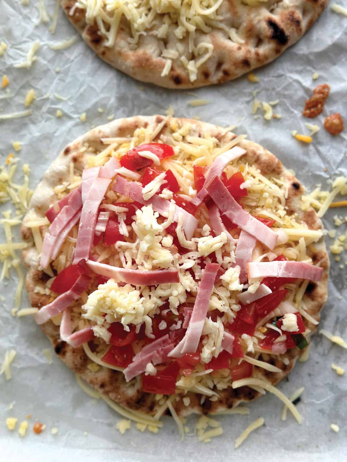 A pita loaded with cheddar cheese, bacon strips and diced tomatoes on parchment paper.