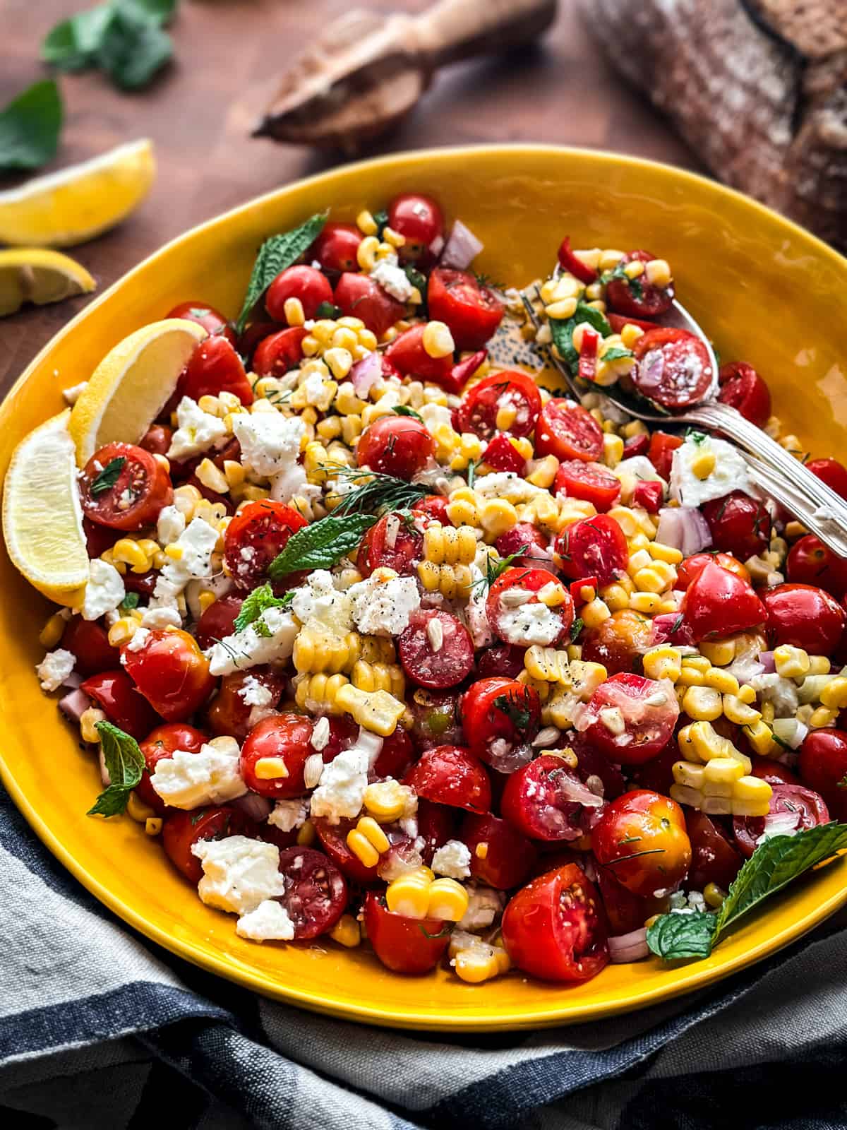 A yellow serving bowl with corn tomato salad with feta, lemon wedges and serving utensils, at the back a citrus juicer and a loaf of bread.