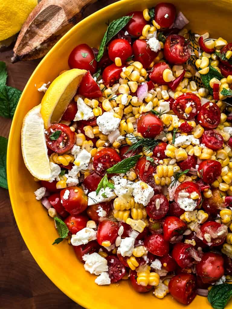A yellow serving bowl with corn tomato salad with feta, lemon wedges and serving utensils.
