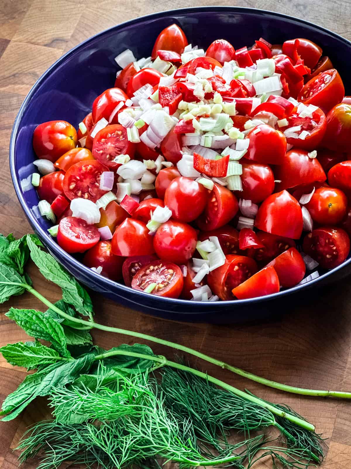 A blue plate with halved cherry tomatoes, sliced chili pepper, chopped onions. Infront of it spearmint leaves and dill. All on a wooden table.