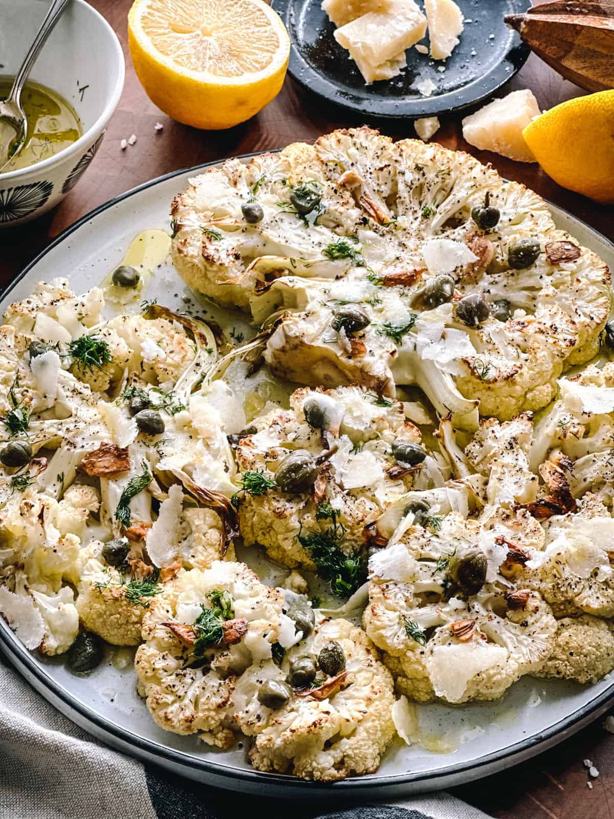 A plate with roasted cauliflower steaks with dill, capers and parmesan cheese. Partial view of two lemon halves, a plate with cheese, a citrus juicer and a cloth napkin.