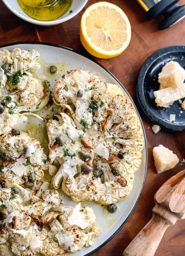 A plate with roasted cauliflower steaks with dill, capers and parmesan cheese. Partial view of two lemon halves, a plate with cheese, a citrus juicer and a cloth napkin.