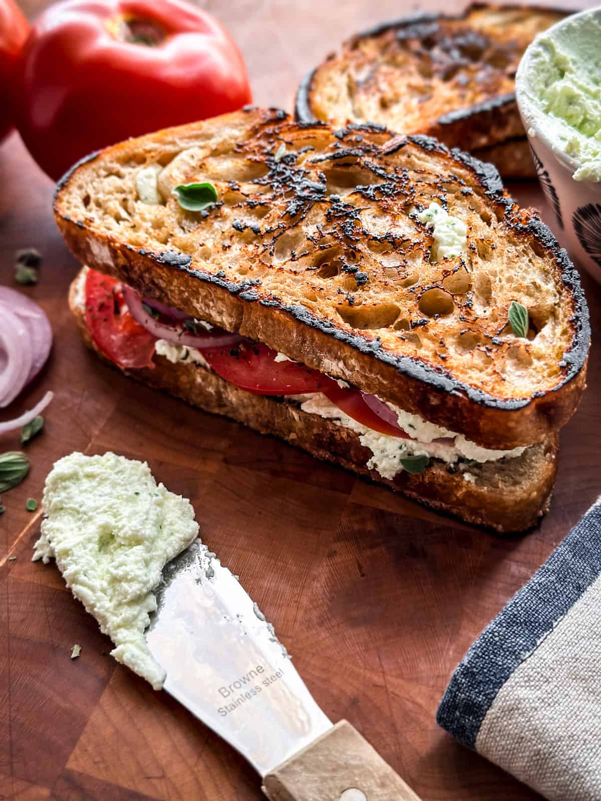 A tomato sandwich on a table. A knife with cheese and a napkin. At the far back a tomato.