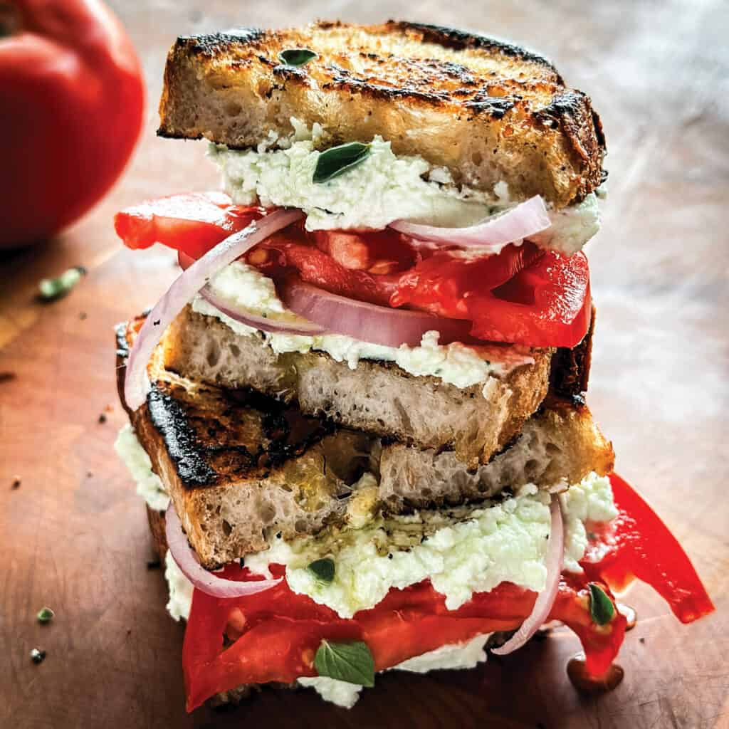 Two tomato sandwich pieces stacked on a table. At the far back a tomato.